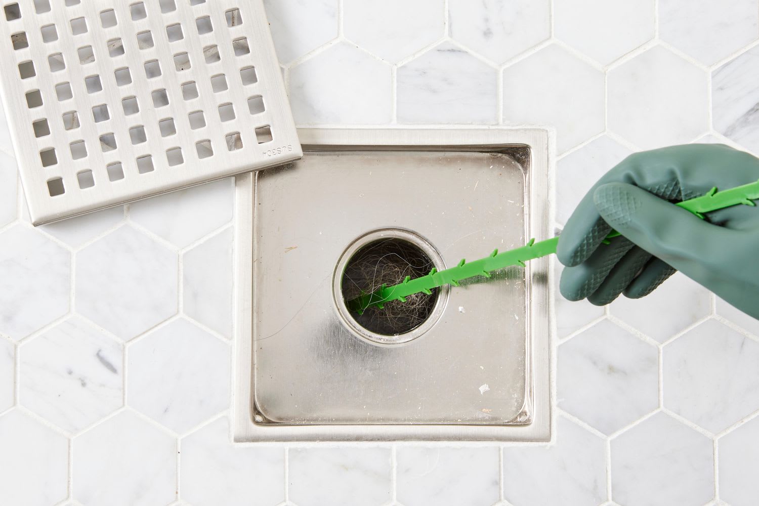 hand in green glove using snake tool to clean shower drain and prevent drain fly infestations from biofilm buildup